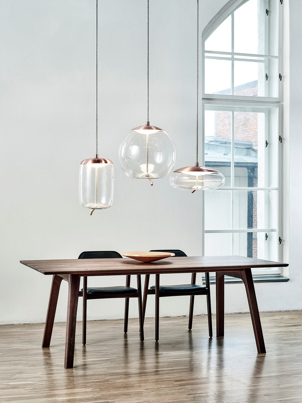 Clear glass Clérachic pendants with copper-toned caps hang above a dark wooden table, with a tall arched window behind.