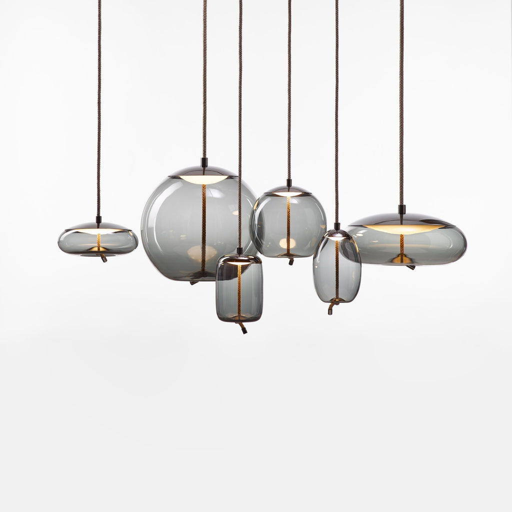 On a white backdrop, six smoke-grey glass Clérachic pendants in round, flat and tall shapes hang at different heights from braided cords.