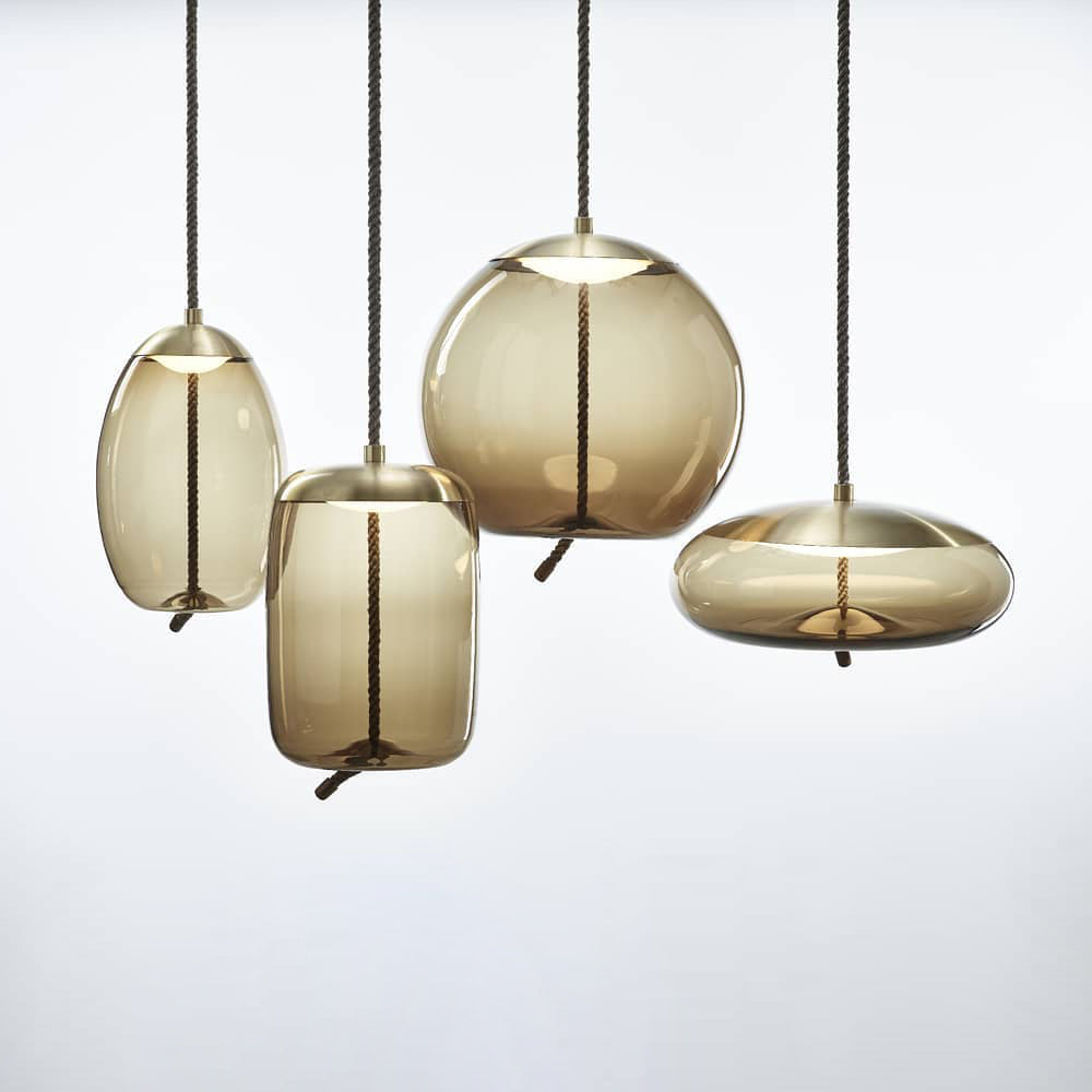 Four amber glass Clérachic pendants with brass-toned caps, in oval, tall, round and flat shapes, hang together on a pale backdrop.