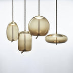 Four amber glass Clérachic pendants with brass-toned caps, in oval, tall, round and flat shapes, hang together on a pale backdrop.