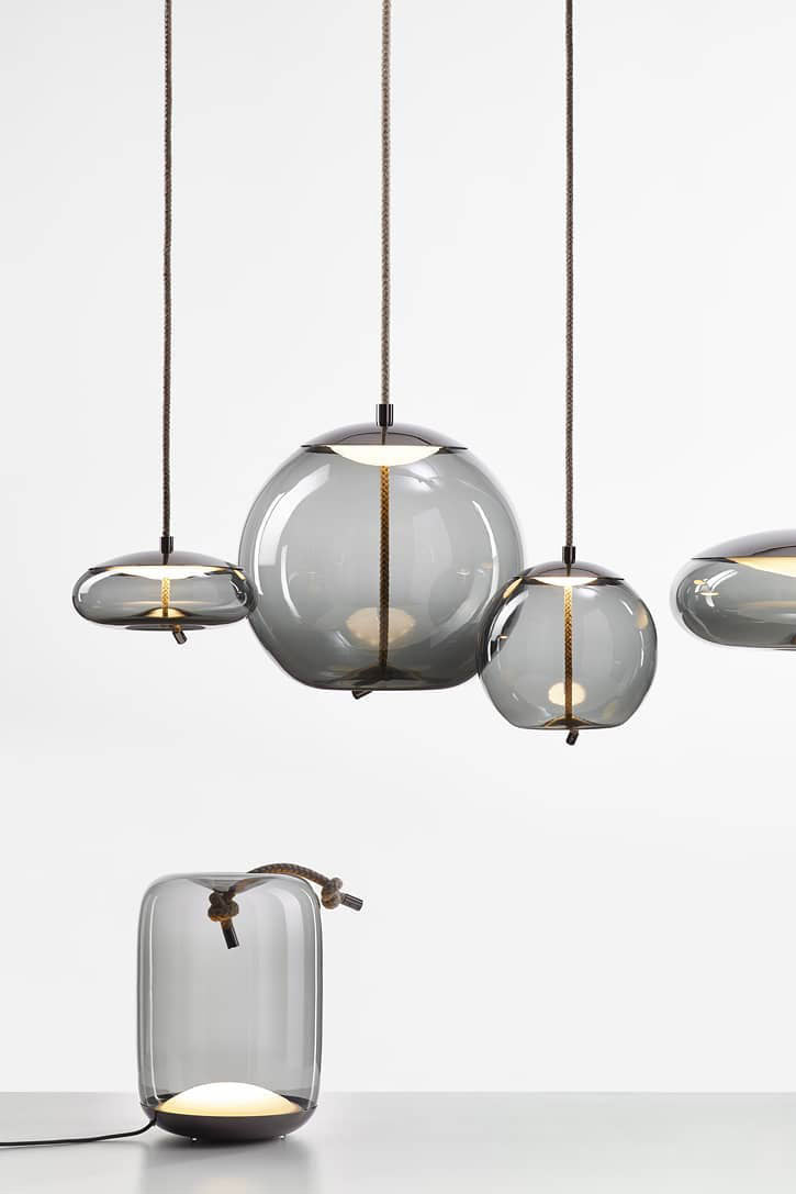 Smoke-grey glass Clérachic pendants hang in a group above a small matching table lamp with a rope handle, on a pale backdrop.