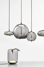 Smoke-grey glass Clérachic pendants hang in a group above a small matching table lamp with a rope handle, on a pale backdrop.