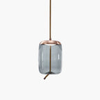 A tall, rounded-cylinder smoke-grey glass Clérachic pendant with a copper-toned cap hangs from a rope cord on a pale backdrop.