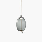 A narrow oval smoke-grey glass Clérachic pendant with a dark cap hangs from a brown braided cord on a pale backdrop.
