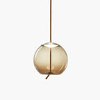 A round amber glass Clérachic pendant with a brass-toned cap and a short rope tail hangs on a pale backdrop.