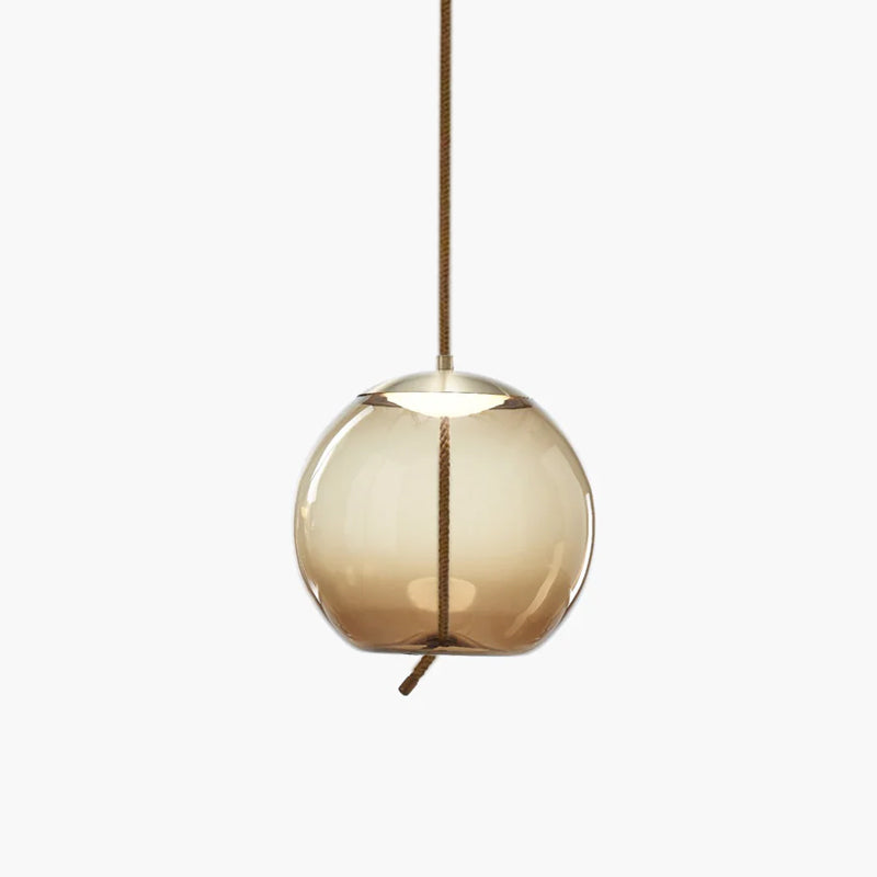 A round amber glass Clérachic pendant with a brass-toned cap and a short rope tail hangs on a pale backdrop.