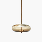 A flat, wide amber glass Clérachic pendant with a brass-toned top and a braided cord hangs on a pale backdrop.