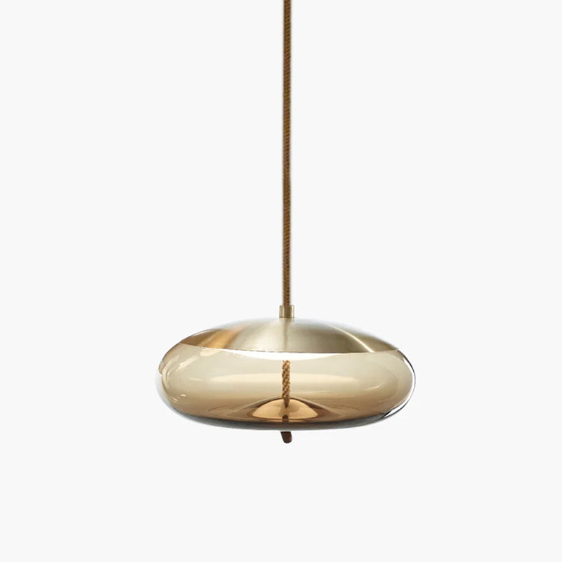 A flat, wide amber glass Clérachic pendant with a brass-toned top and a braided cord hangs on a pale backdrop.