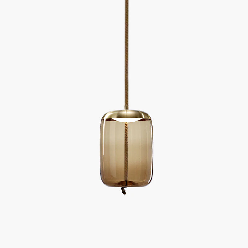 A tall amber glass Clérachic pendant with a brass-toned cap hangs from a brown braided cord on a pale backdrop.