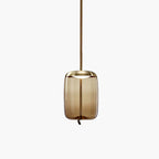 A tall amber glass Clérachic pendant with a brass-toned cap hangs from a brown braided cord on a pale backdrop.