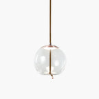 A round clear glass Clérachic pendant with a copper-toned cap hangs from a braided cord on a pale backdrop.