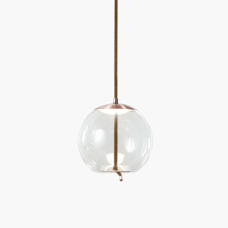 A round clear glass Clérachic pendant with a copper-toned cap hangs from a braided cord on a pale backdrop.