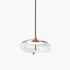 A flat, wide clear glass Clérachic pendant with a copper-toned top hangs from a braided cord on a pale backdrop.