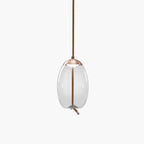 A narrow oval clear glass Clérachic pendant with a copper-toned cap hangs from a brown braided cord on a pale backdrop.