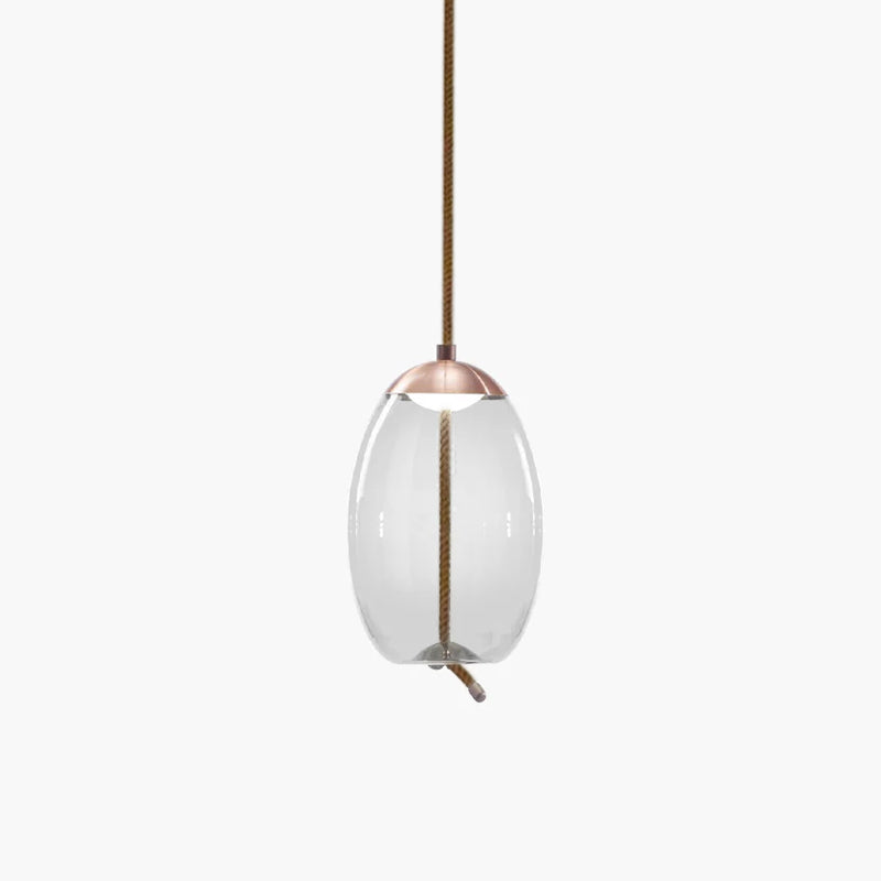 A narrow oval clear glass Clérachic pendant with a copper-toned cap hangs from a brown braided cord on a pale backdrop.
