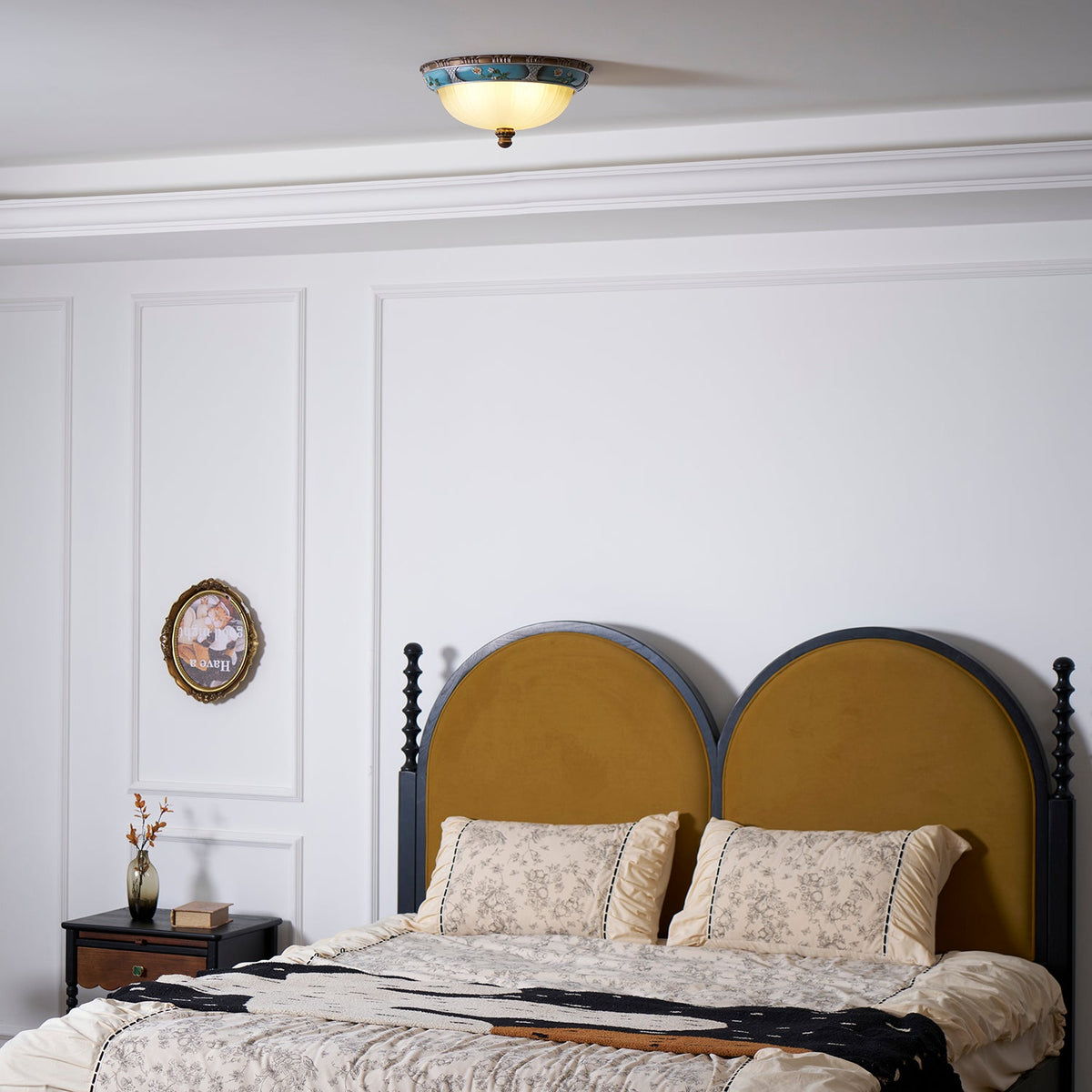 Clérachic ceiling light seen from the foot of a bed, high above a mustard arched headboard and a white panelled wall.