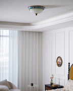Clérachic ceiling light above a white bedroom with floor-length curtains, a panelled wall, an oval picture and a bedside table.