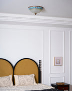 Clérachic ceiling light seen from below beyond a white cornice, above an arched mustard headboard and a floral cushion.