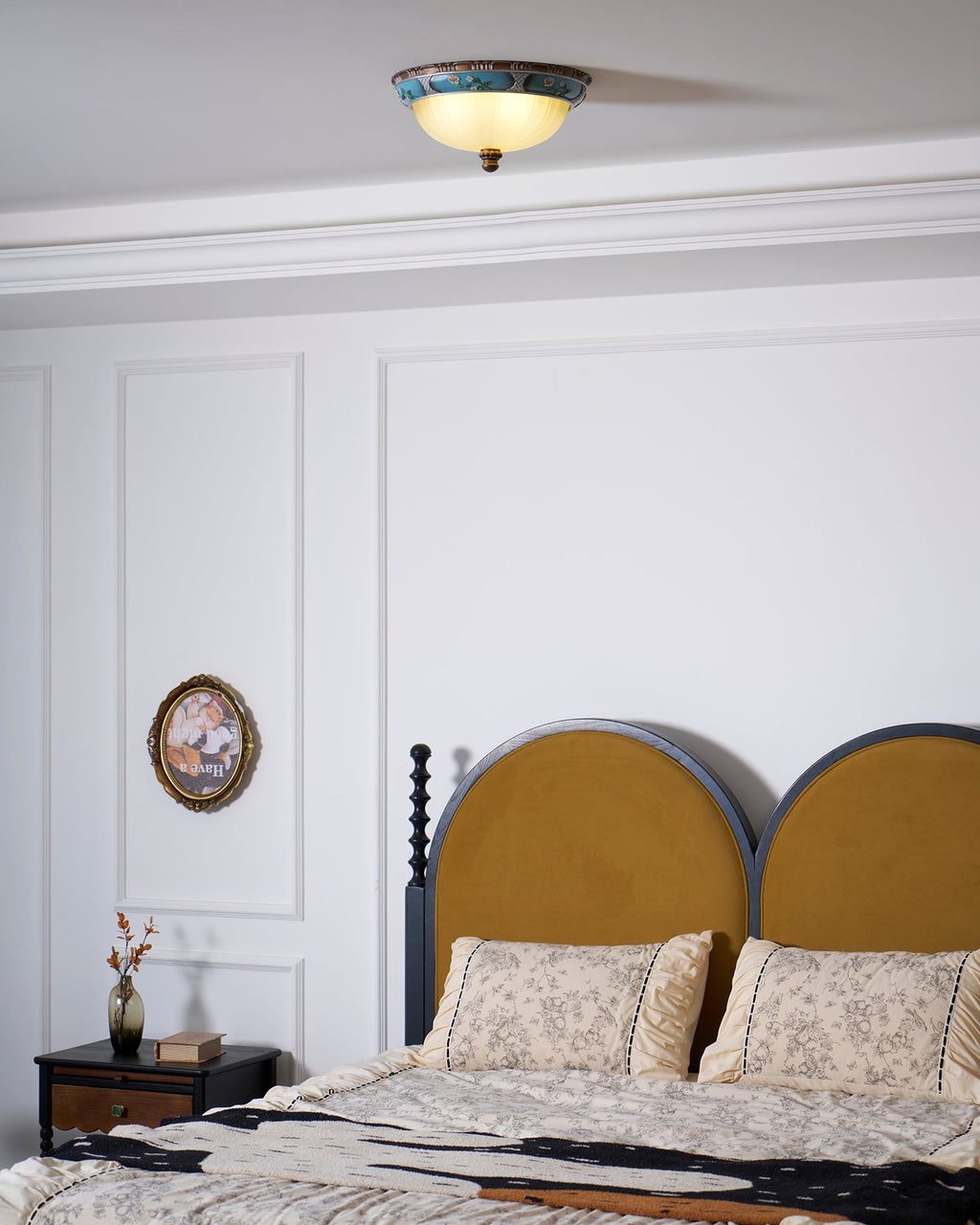 Clérachic ceiling light above a bedroom with a mustard arched headboard, a floral bedspread and a panelled white wall.