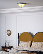 Clérachic ceiling light above a bedroom with a mustard arched headboard, a floral bedspread and a panelled white wall.
