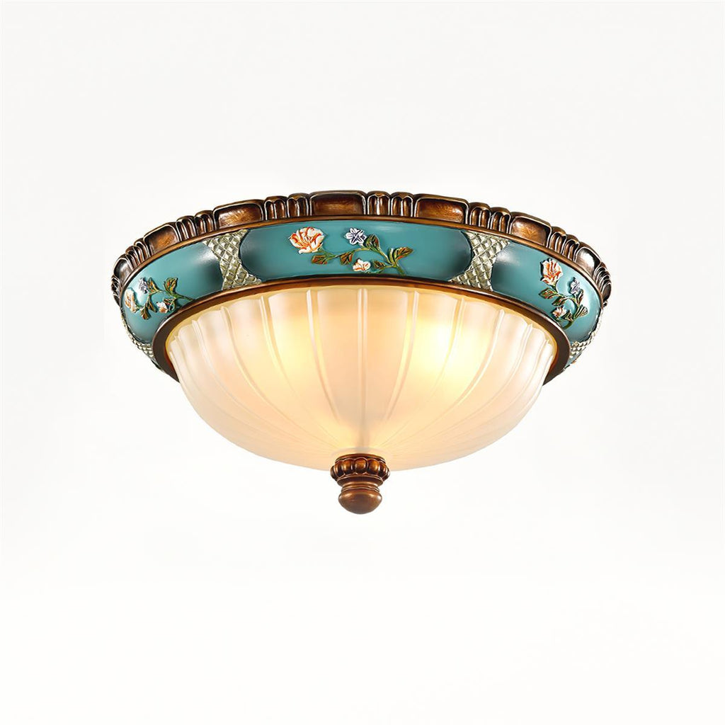 Lit Clérachic ceiling light on a white backdrop, the ribbed glass glowing warm under a turquoise band of painted flowers.