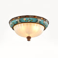Lit Clérachic ceiling light on a white backdrop, the ribbed glass glowing warm under a turquoise band of painted flowers.