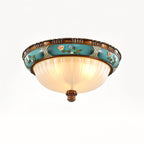 Lit Clérachic ceiling light on a white backdrop, the ribbed glass glowing warm under a turquoise band of painted flowers.