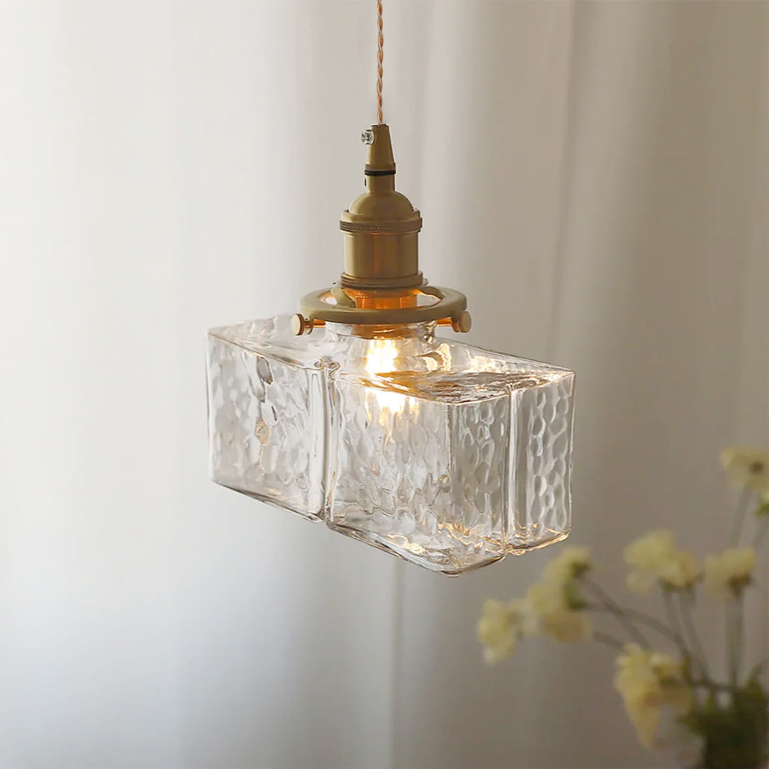Close-up of a lit Clérachic boxy hammered-glass pendant light, Model B, angled beside sheer curtains and yellow wildflowers.
