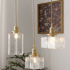 All three Clérachic boxy hammered-glass pendant light shapes lit near a gold-framed floral print and a potted fern.