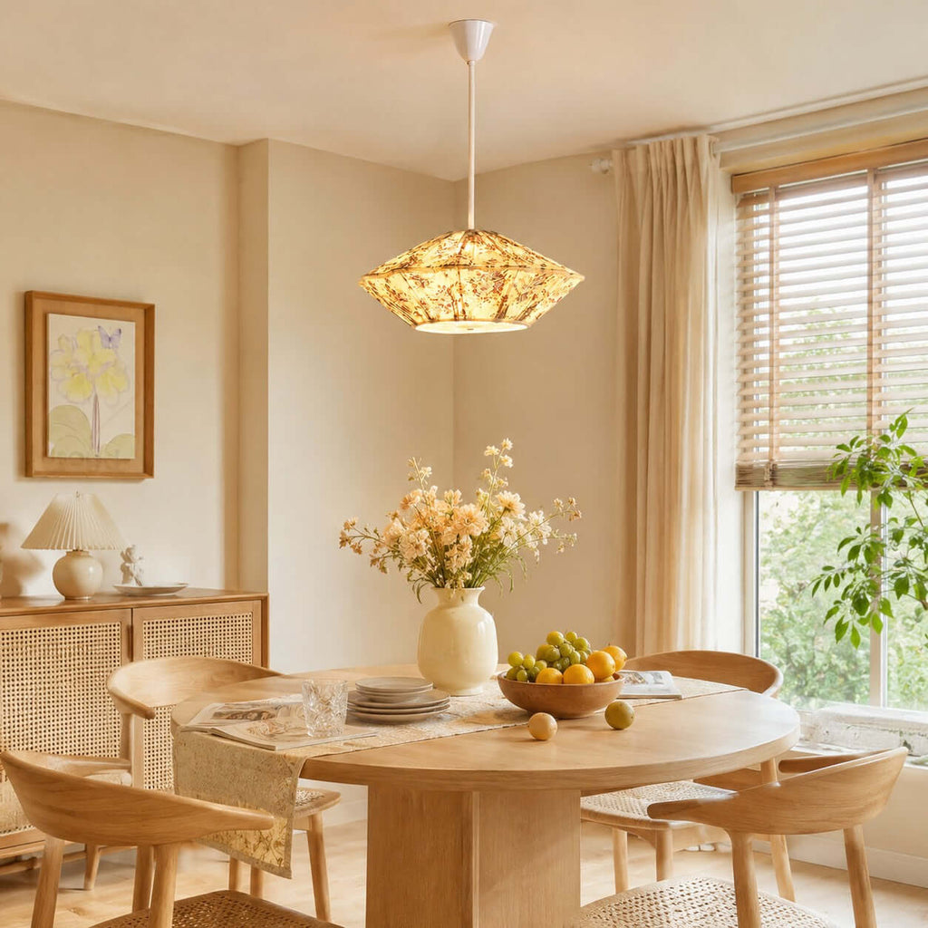 A glowing Clérachic pendant with a floral pleated shade hangs above a round oak table with a bowl of lemons and a vase of flowers.