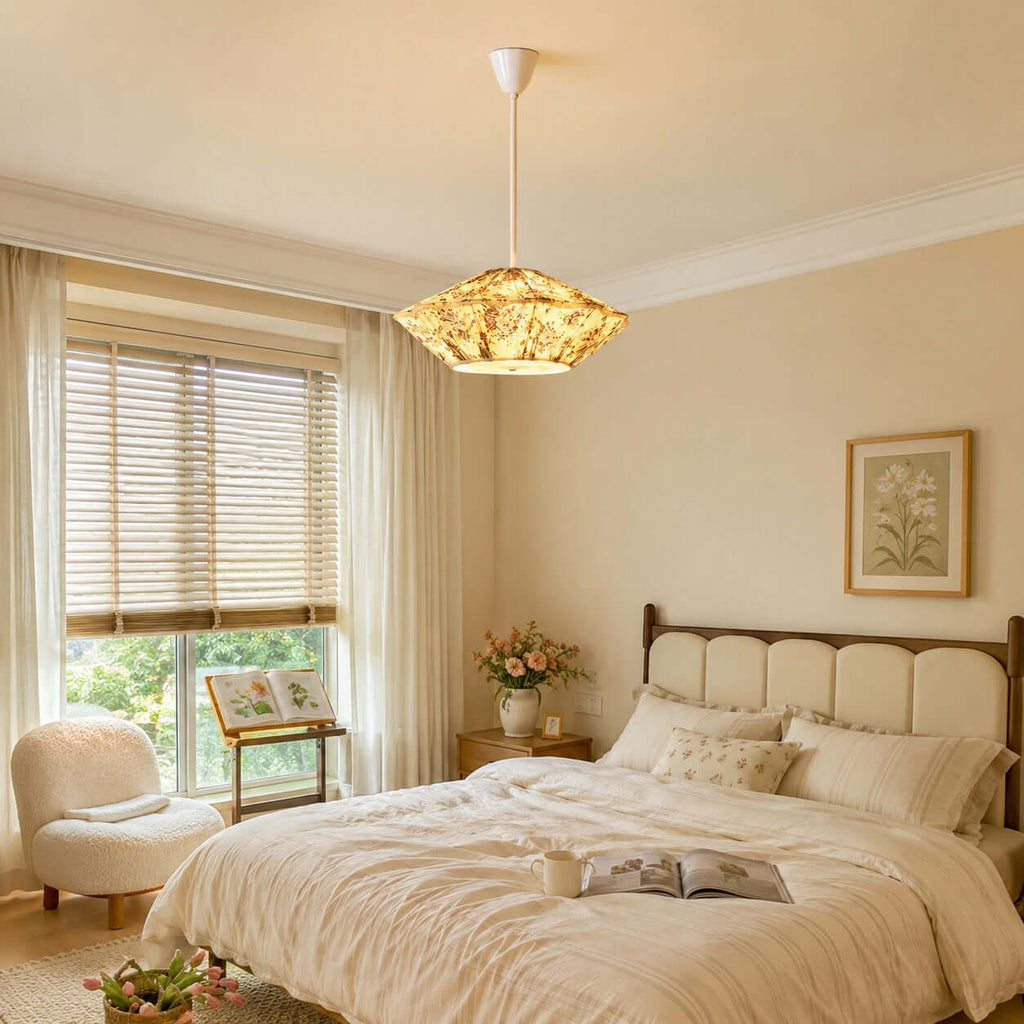 A Clérachic pendant with a lit floral shade hangs in a bedroom with a cream upholstered bed and slatted window blinds.