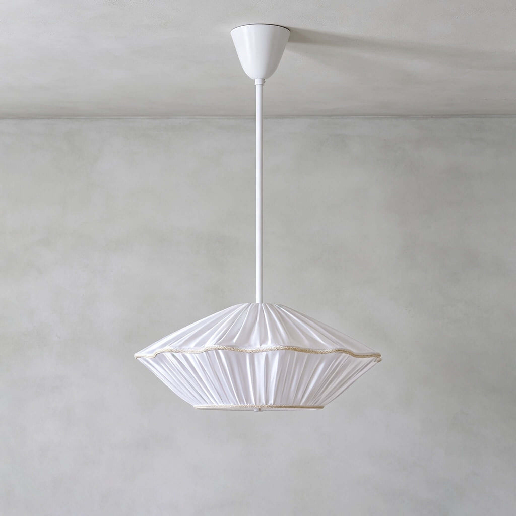 This Clérachic pendant has a white pleated shade with a thin gold-toned trim, hanging from a white rod on a grey wall.