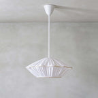 This Clérachic pendant has a white pleated shade with a thin gold-toned trim, hanging from a white rod on a grey wall.