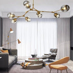 Clérachic chandelier in gold and smoke glass, its arms branching over a living room of sheer curtains and a lounge chair.