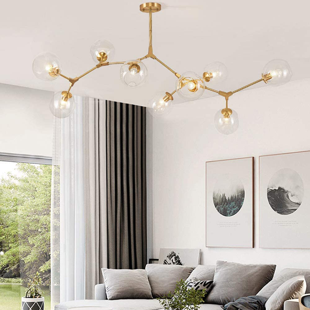 Clear globes on the gold Clérachic chandelier at its widest, branching across a living-room ceiling beside a garden window.