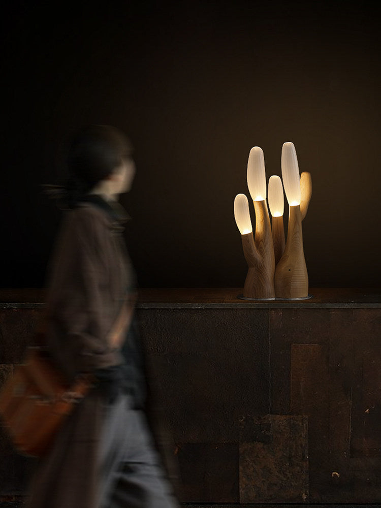 In a dim room, this light-wood Clérachic table lamp sits on a stone counter as a figure walks past.