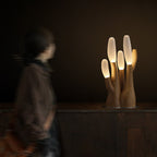 A silhouette drifts past this light-wood Clérachic table lamp, lit on a dark textured counter.