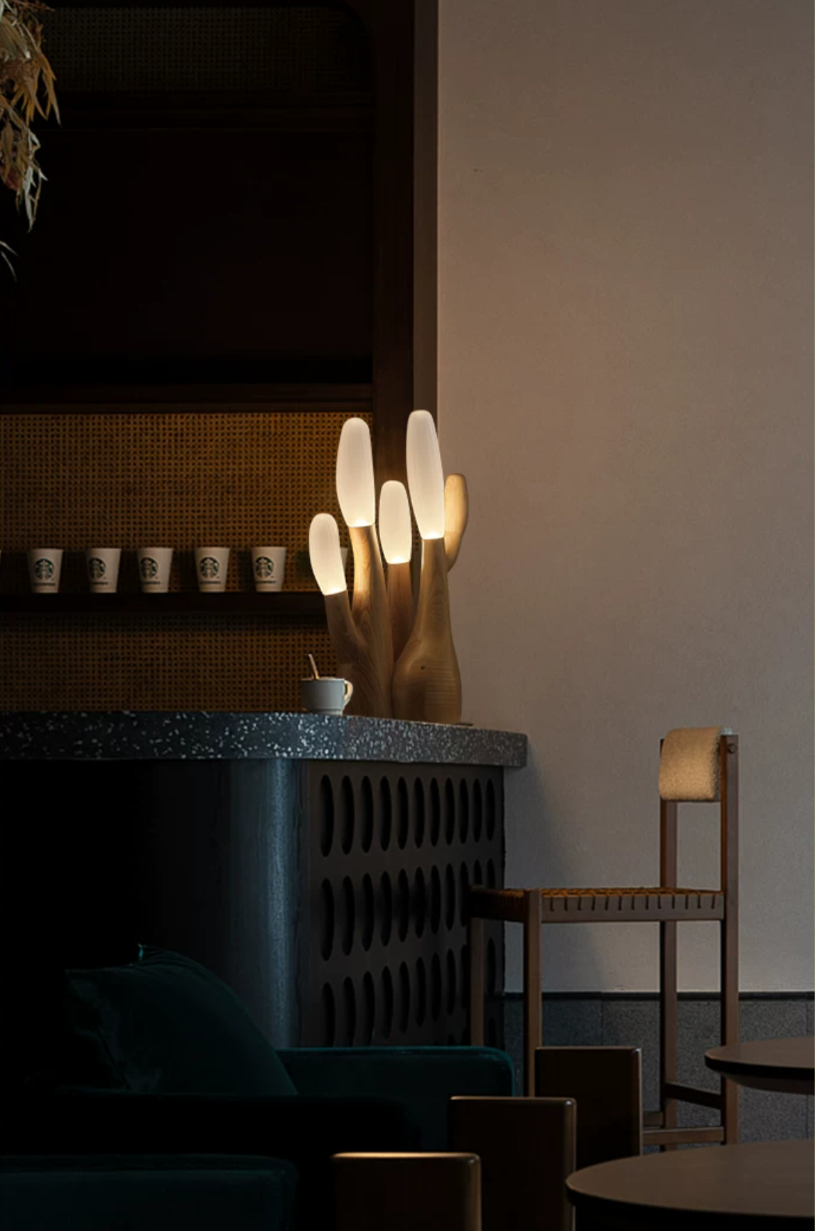 This black Clérachic table lamp glows on a dark counter in a café corner with cups and a chair.