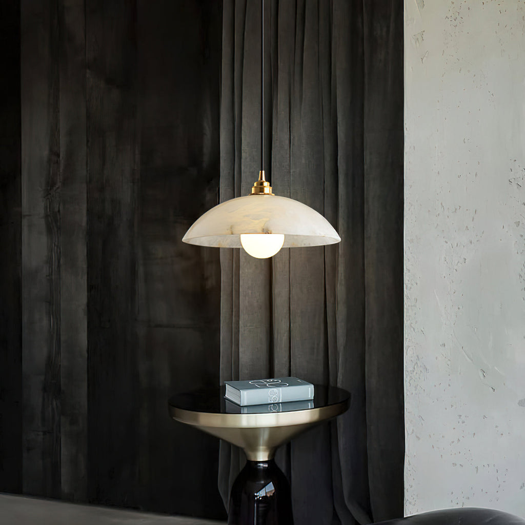 Alabaster dome pendant from Clérachic in front of dark textured drapes, above a brass-topped side table holding a stack of books.