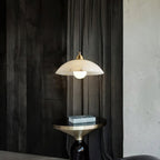 Alabaster dome pendant from Clérachic in front of dark textured drapes, above a brass-topped side table holding a stack of books.