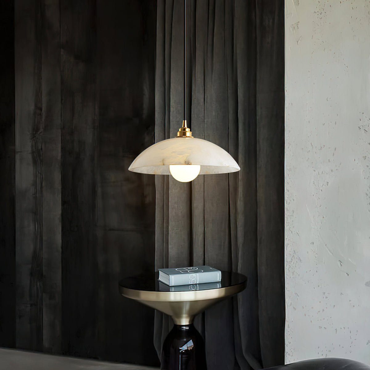 Alabaster dome pendant from Clérachic in front of dark textured drapes, above a brass-topped side table holding a stack of books.