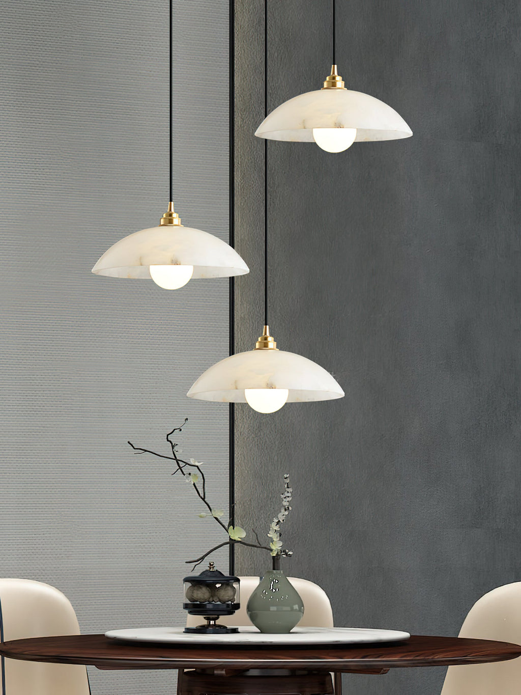 Three Clérachic alabaster domes at different heights over a round dining table with a vase and a bare branch, against a grey wall.