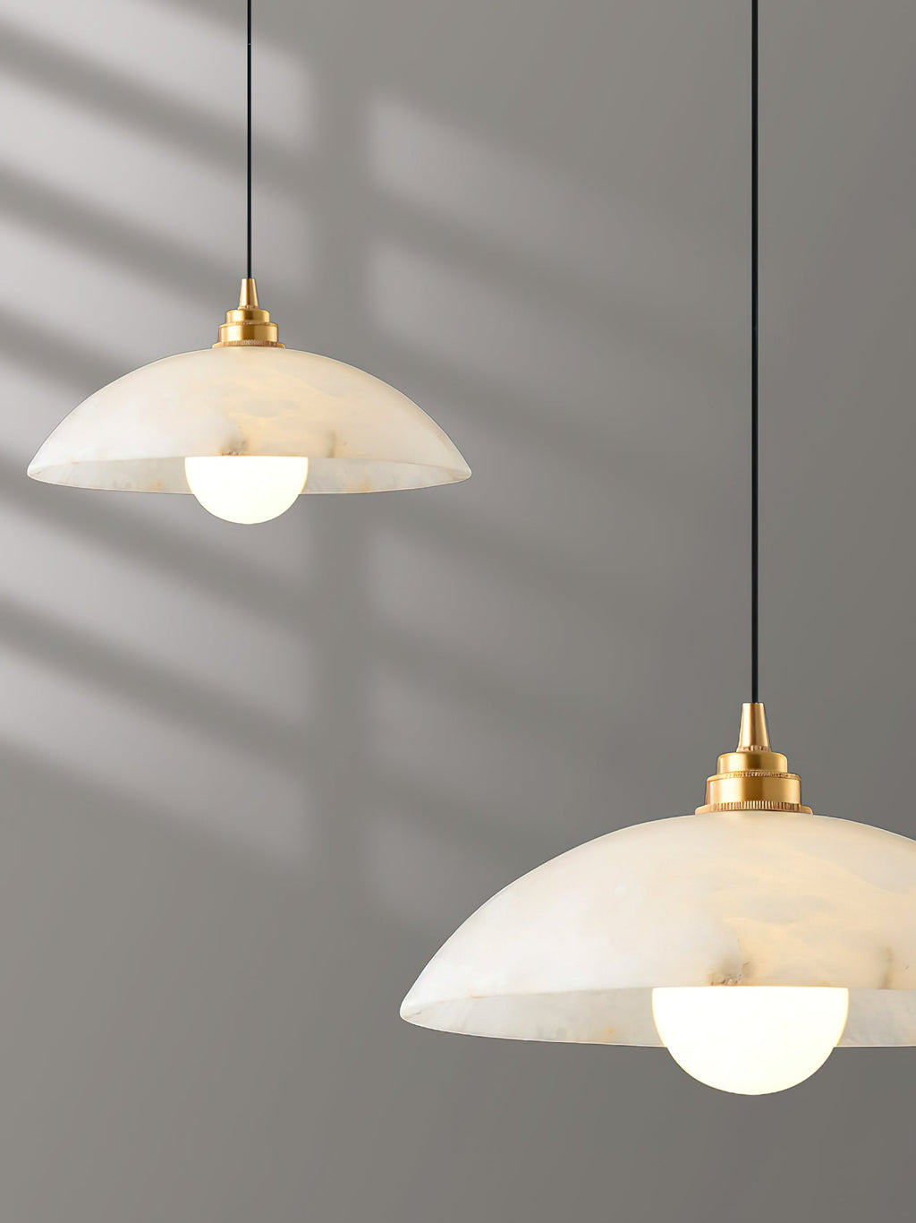 Two alabaster domes of this Clérachic pendant at different heights, in cropped close view against a grey wall striped with sunlight.
