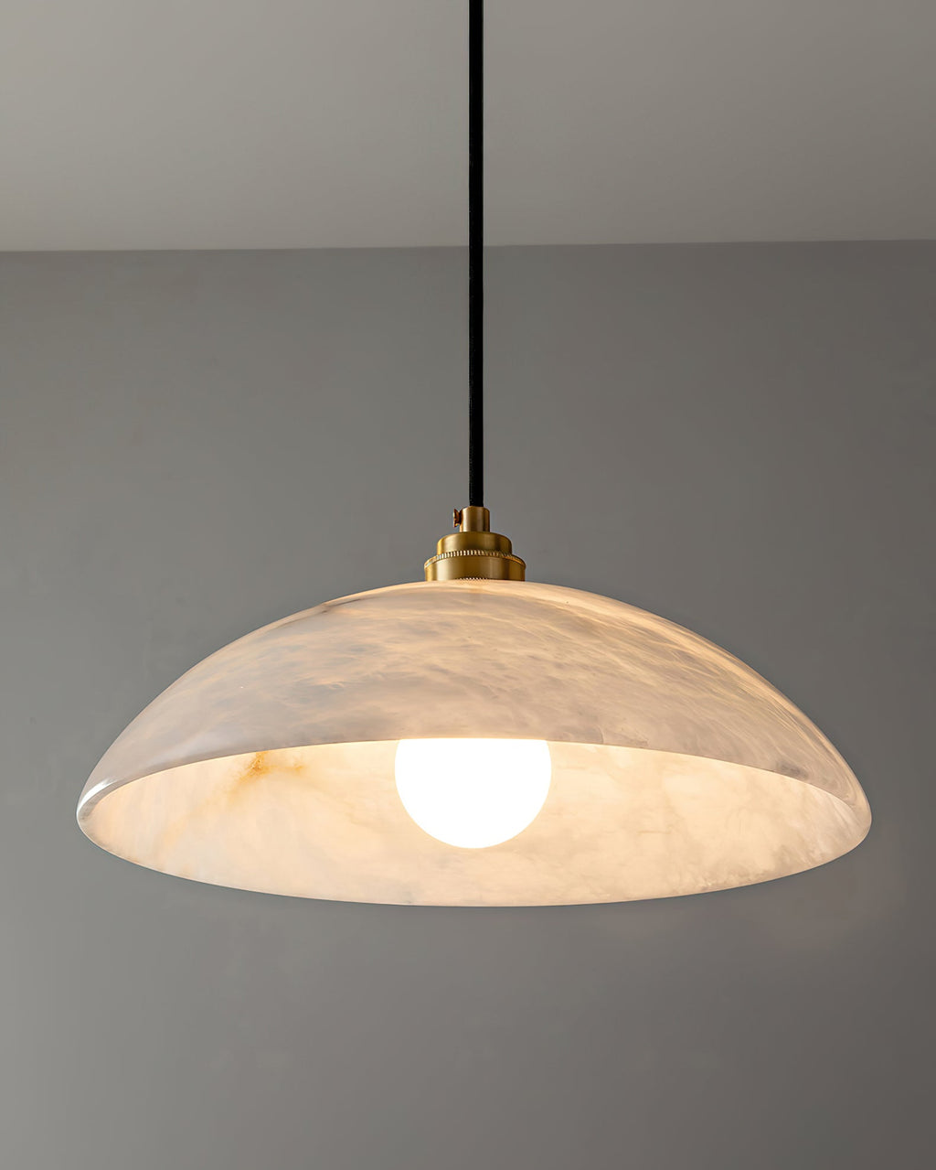 Lit Clérachic alabaster pendant with a bright bulb inside, so the stone's grey veining shows through the dome against a grey wall.