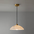 A slightly larger view of this Clérachic pendant, with the brass ceiling plate, black cord and warm-lit alabaster dome against a grey wall.
