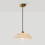 Clérachic alabaster pendant hung on a long black cord from its brass ceiling plate, lit warm, against a light grey wall.