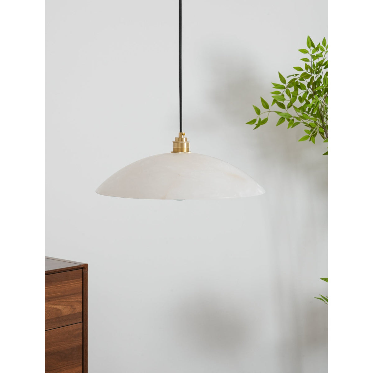 Wide, shallow alabaster dome of a Clérachic pendant seen from below, with a leafy plant and a walnut cabinet beside it.