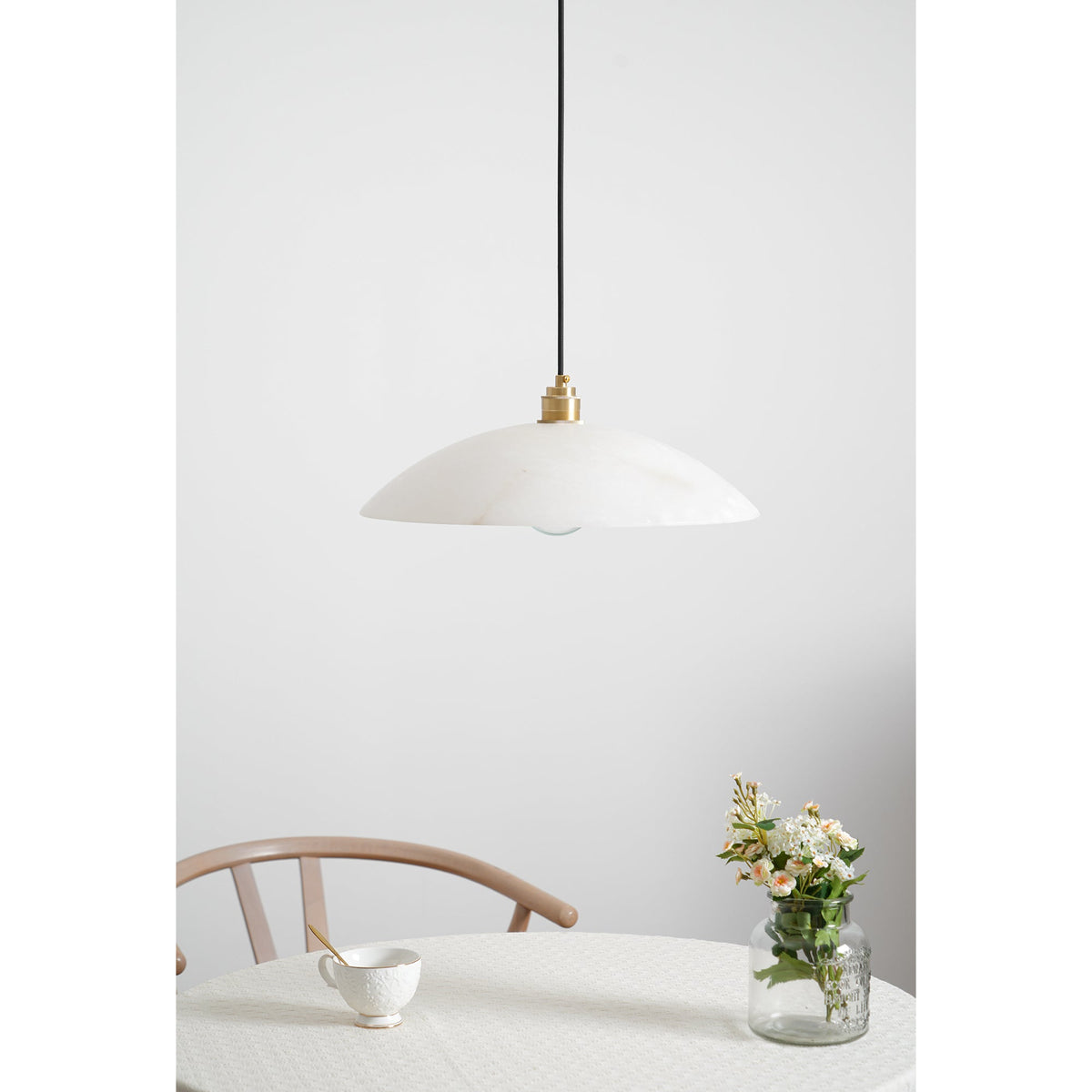 Clérachic alabaster pendant above a round table, with a wishbone chair, a white cup and a vase of flowers on a pale wall.