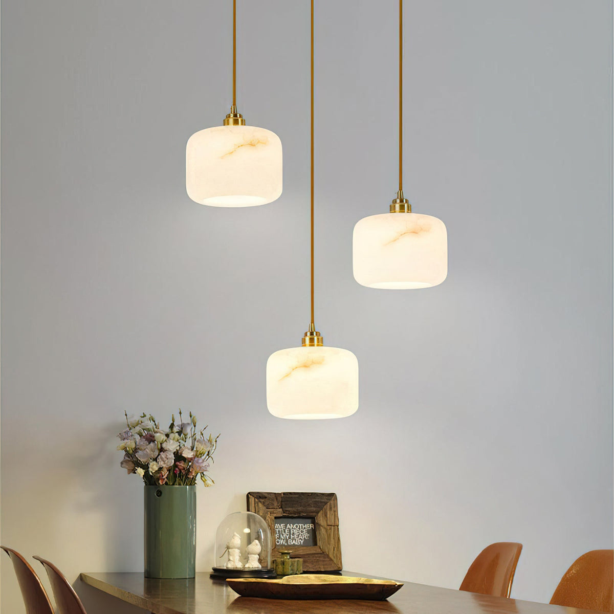 A trio of Clérachic alabaster pendants on brass rods above a dark wood table, with a vase of flowers and framed art below.