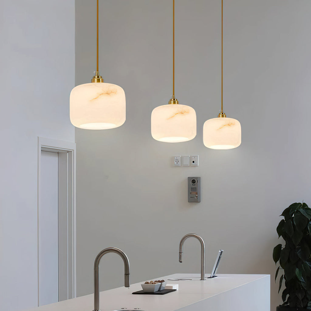 Three Clérachic alabaster pendants in graduated sizes glowing in a kitchen, above a white island with two arched taps.
